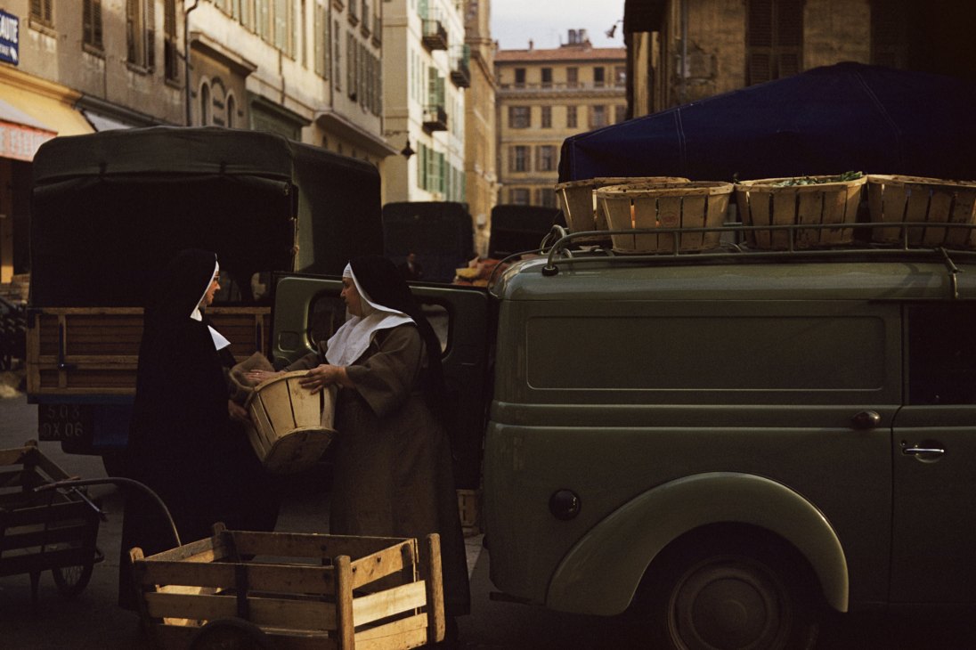 Two nuns in white habits loading wooden crates onto a vintage truck in a European city street