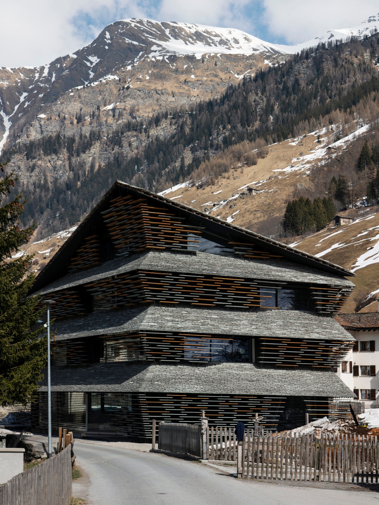 Modern wooden-slat building with stone facade nestled in snow-covered Alpine valley with forested mountains.