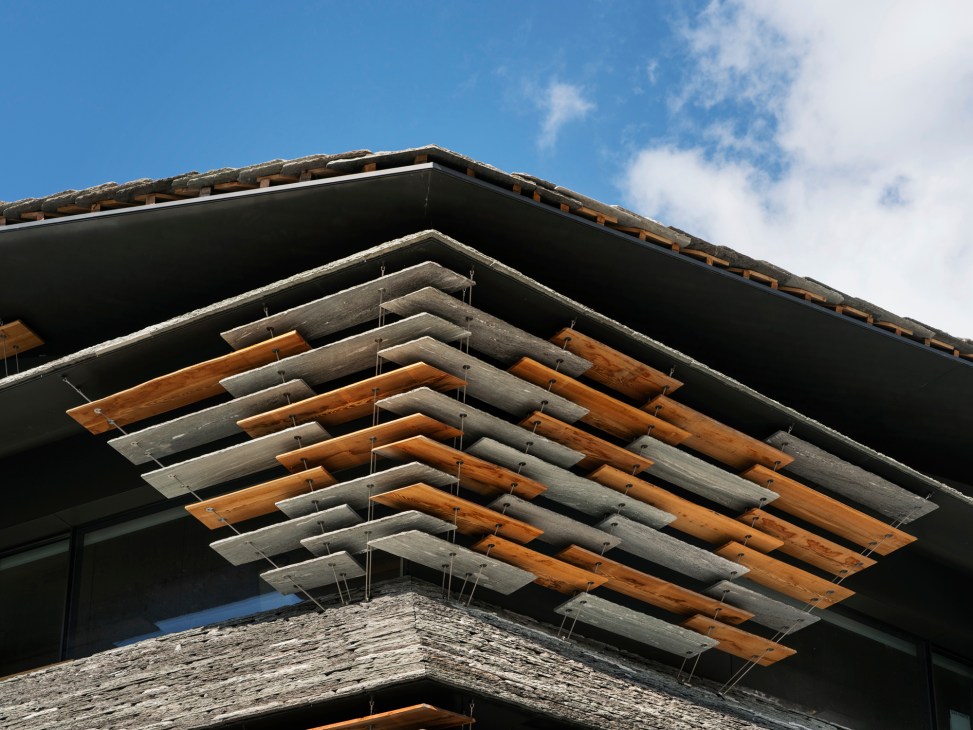 Modern stone building facade with large layered stone slabs that appear to levitate, set in a mountainous alpine landscape.