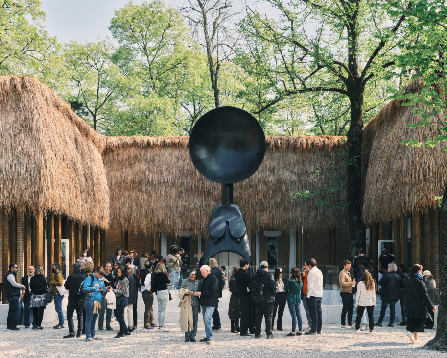 Visitors gather in front of Simone Leigh's US Pavilion featuring a large black spherical sculpture and thatched roof structure at Venice Biennale.
