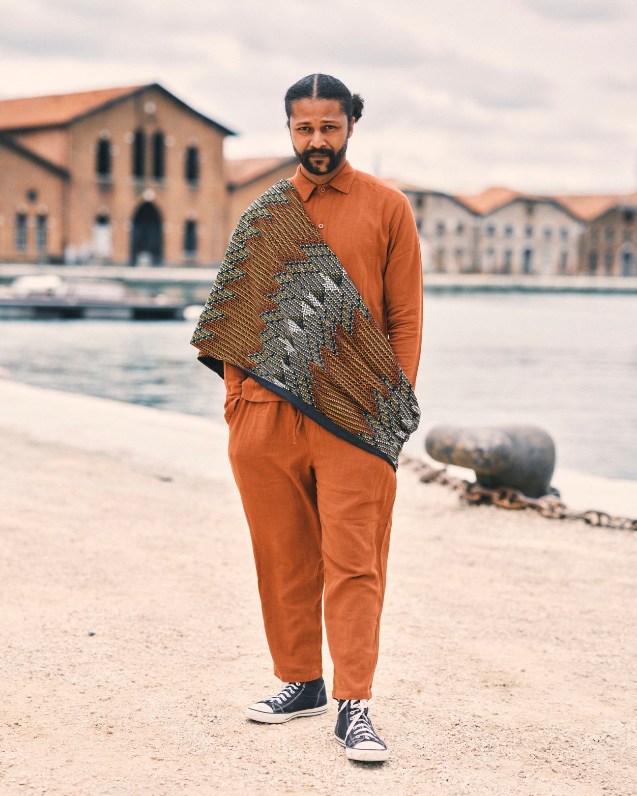 Man in terracotta clothing holding patterned textile at Venice waterfront with historic buildings behind.