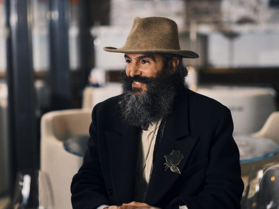 Man with beard and hat in dark jacket sitting at table with drink in elegant interior setting