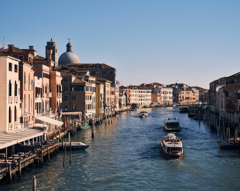 Venice's Grand Canal with historic palazzos lining both sides, boats navigating the waterway under clear blue sky