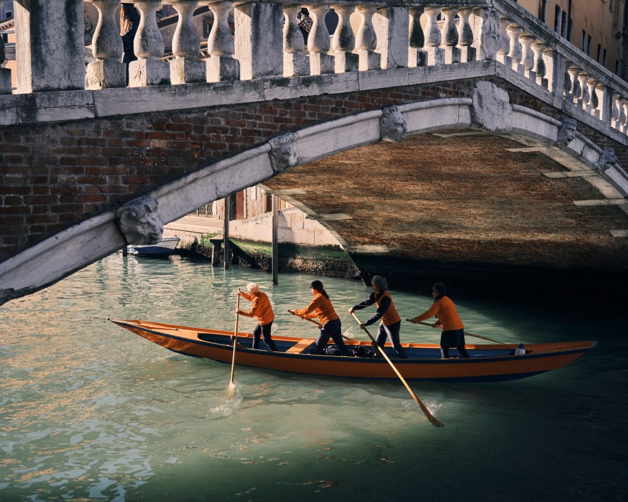 Four rowers in orange shirts practicing traditional Venetian rowing technique in wooden boat under historic bridge