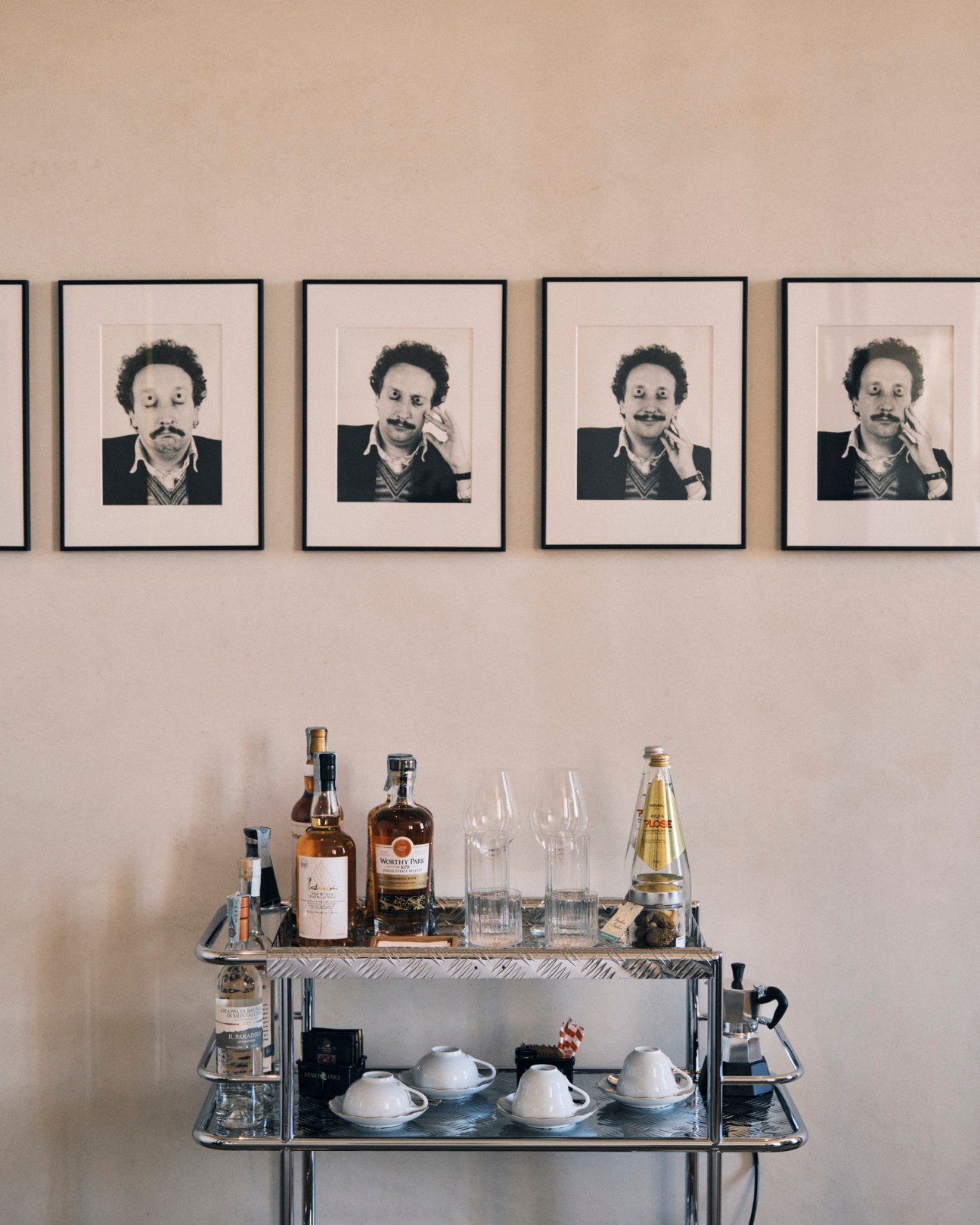 Stylish drinks trolley with bottles, glasses and coffee service on metal cart with framed portraits on wall above