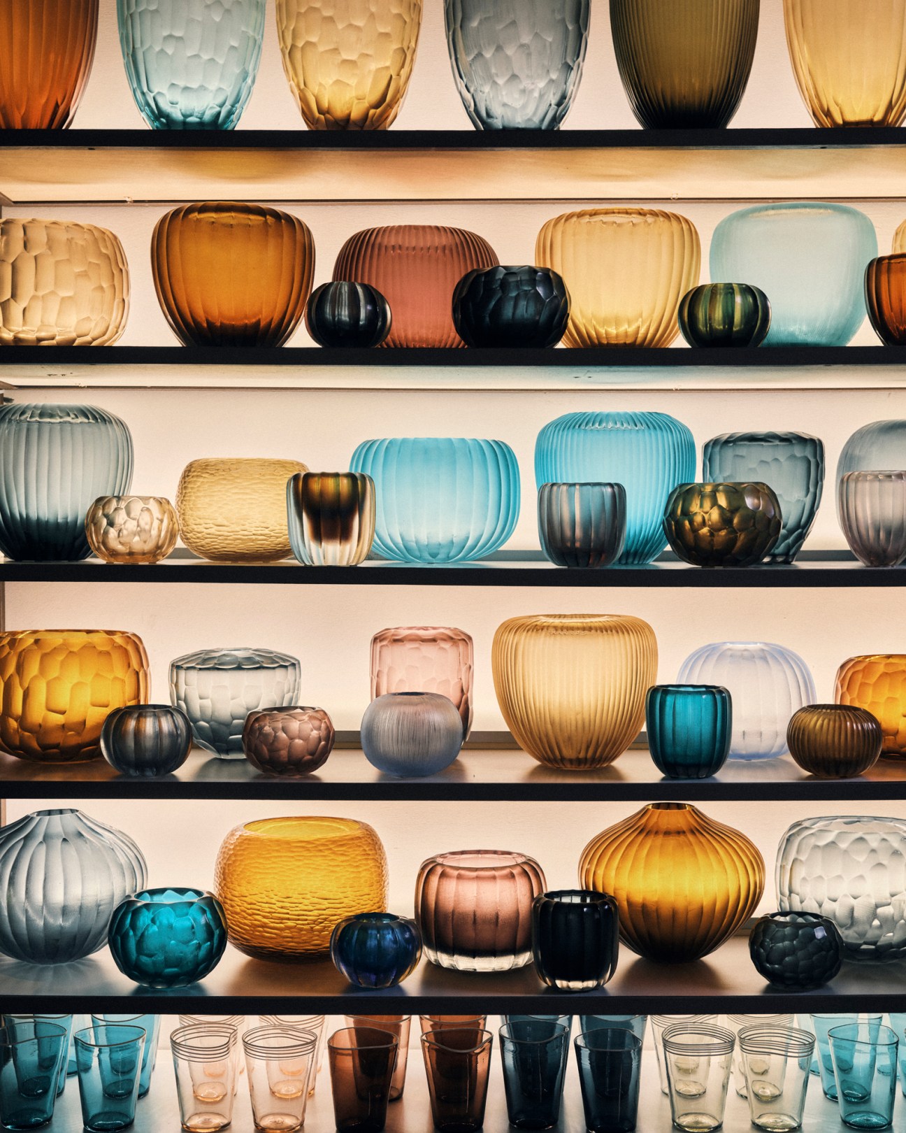 Collection of colorful blown glass vases and vessels displayed on white shelves in various jewel tones
