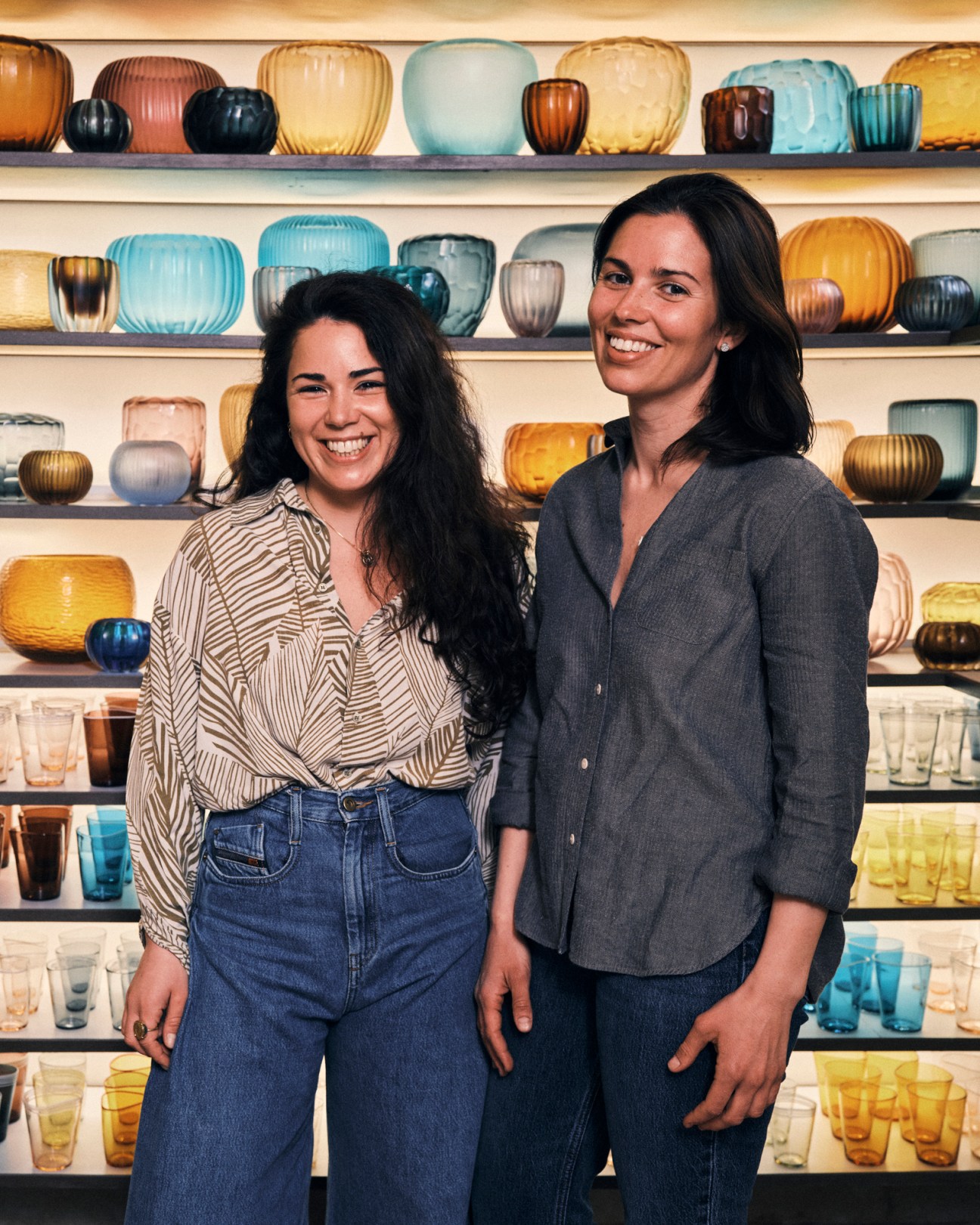 Elena and Margherita Micheluzzi standing in their glass design studio surrounded by colorful blown glass vases and bowls