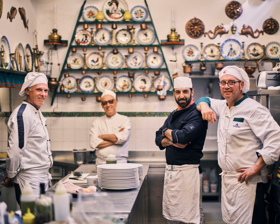 Four chefs in white uniforms and chef hats posing in kitchen of Trattoria da Romano with decorative plates on walls