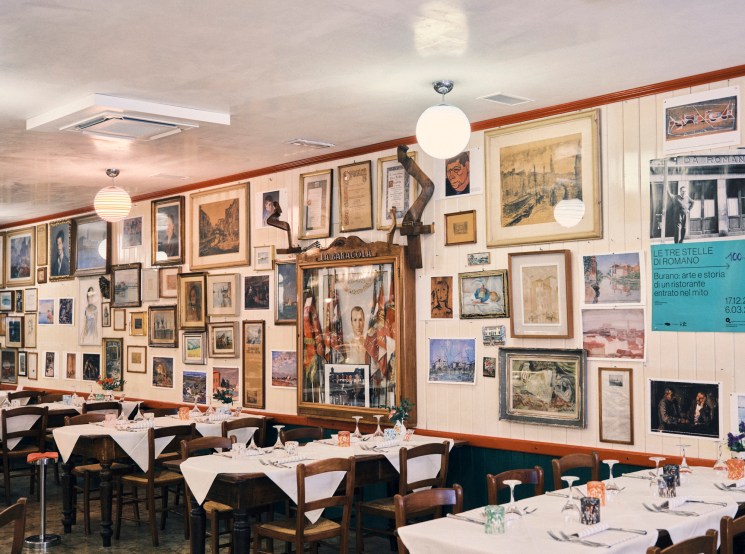 Interior of traditional Italian restaurant with white tablecloths, wooden chairs, and walls covered in framed artwork