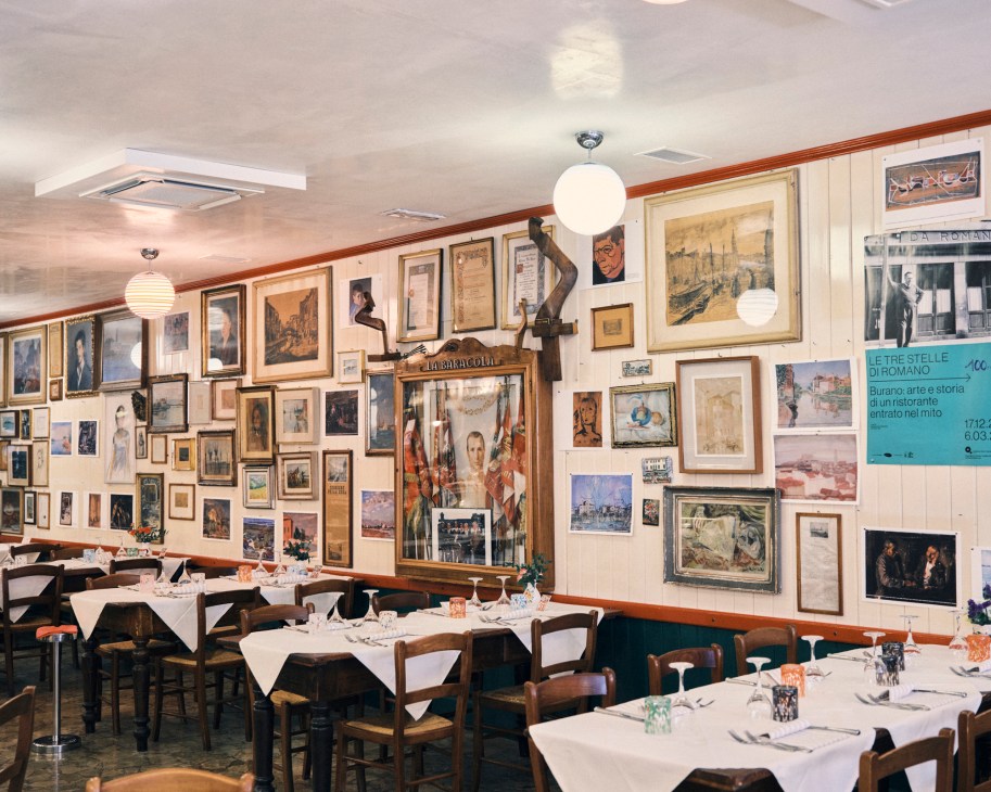 Interior of traditional Italian restaurant with white tablecloths, wooden chairs, and walls covered in framed artwork