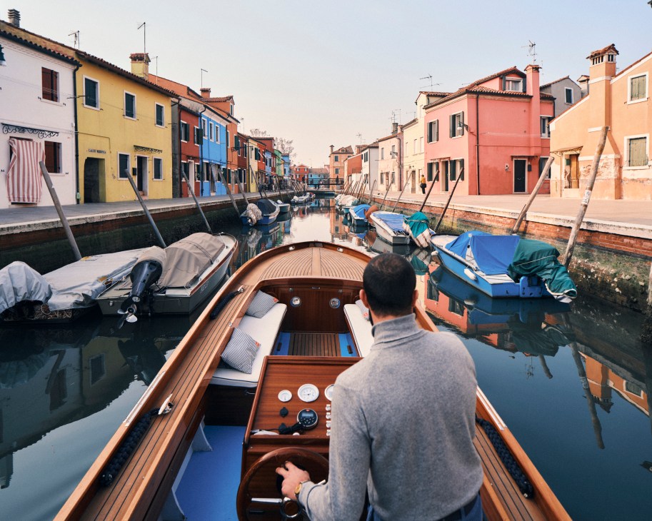 Person navigating wooden boat through narrow canal lined with colorful houses in Burano