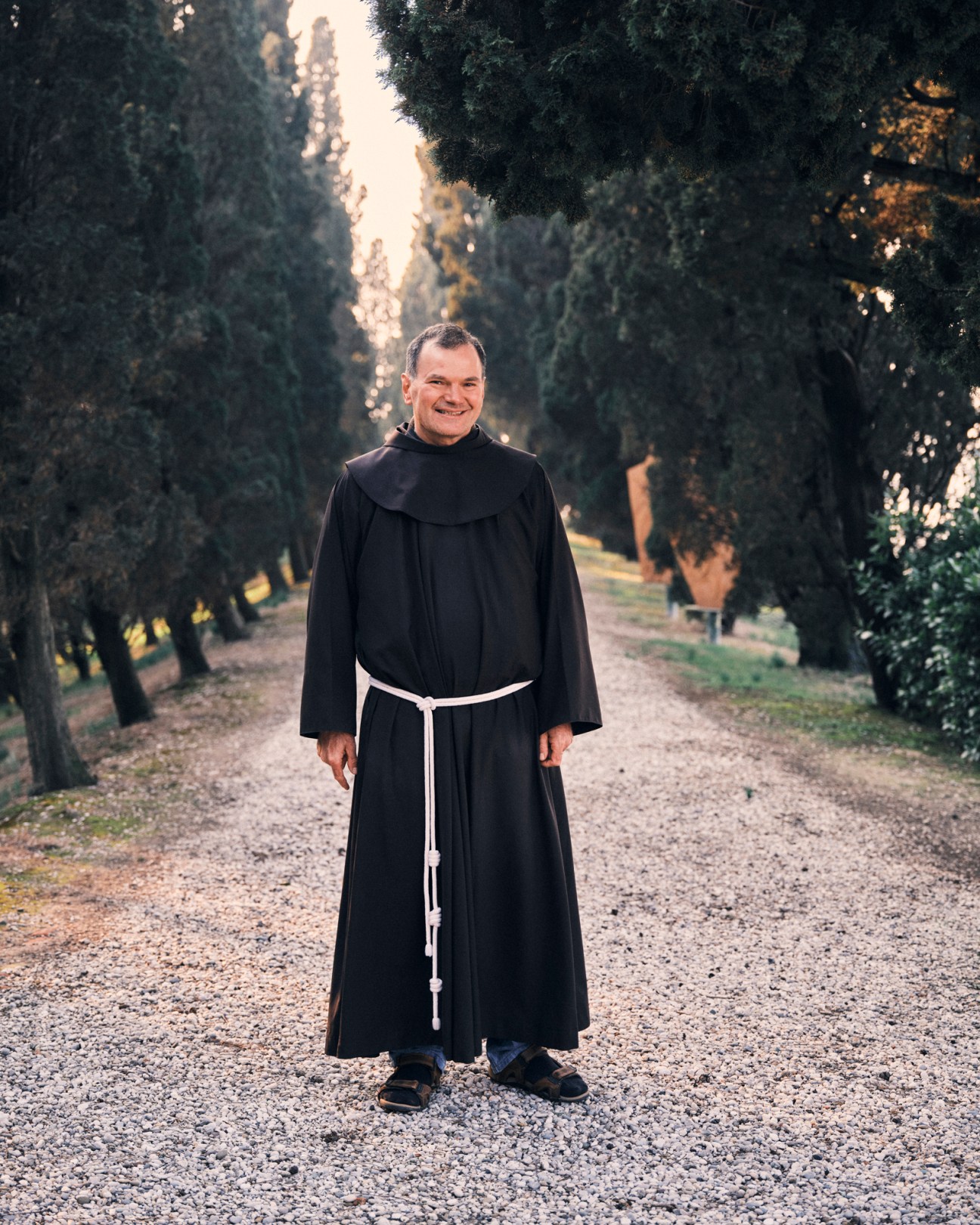 Brother Agostino in brown Franciscan robes standing on tree-lined path at San Francesco del Deserto monastery