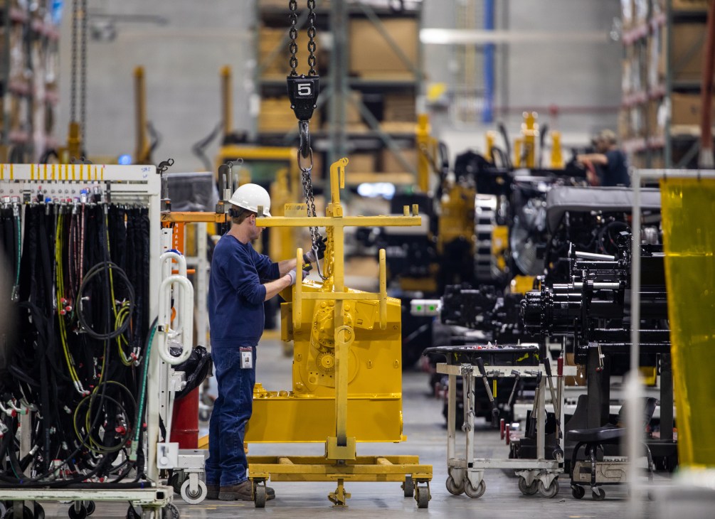 Worker in hard hat assembling yellow industrial machinery with chain hoist in factory.