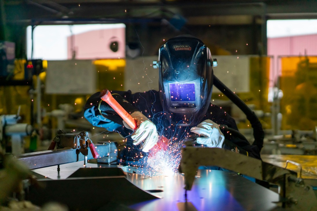 A welder wearing a protective helmet performs welding work on metal, with bright sparks flying in an industrial factory setting.