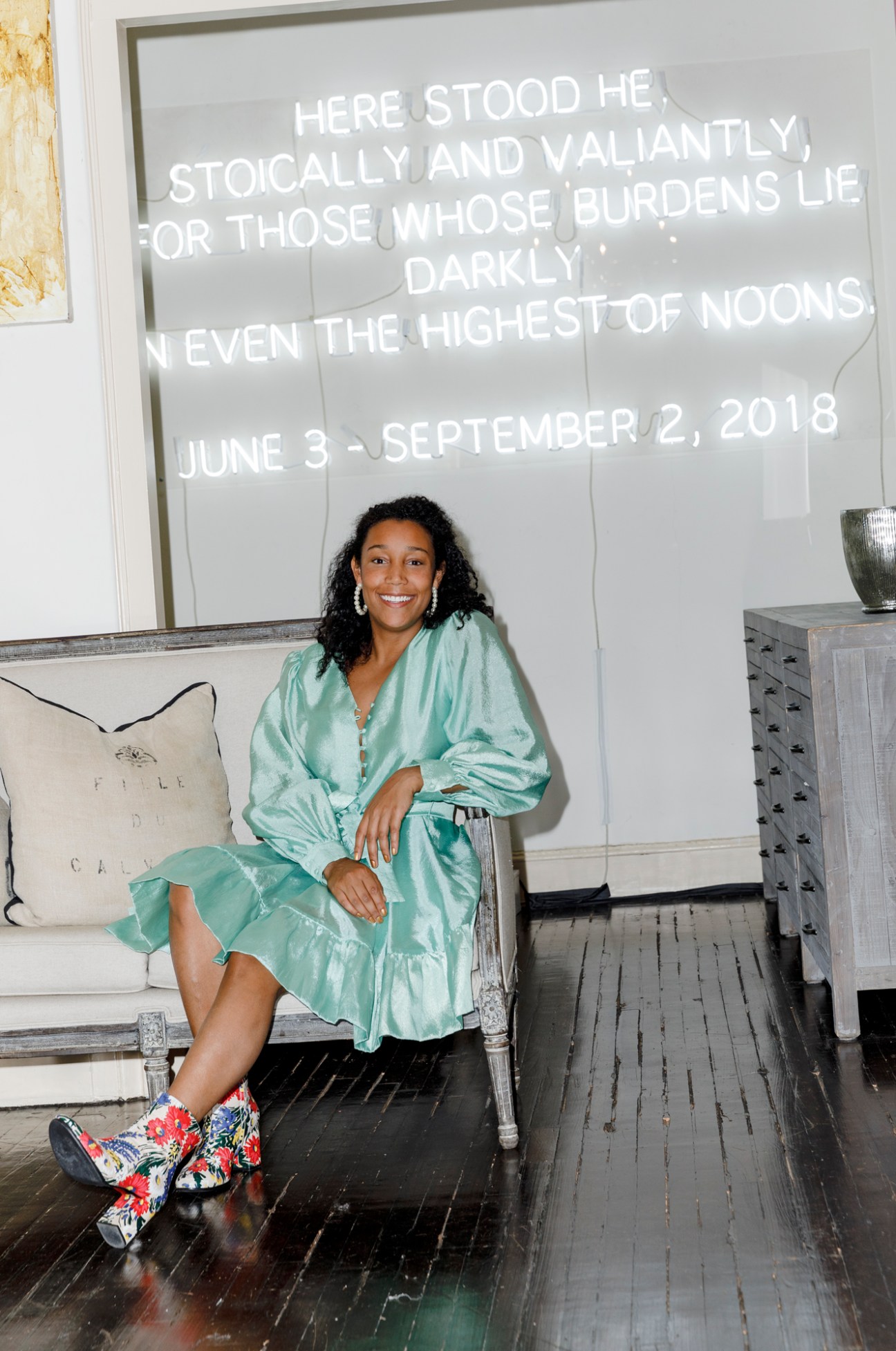 Victoria Rogers sits on a white bench wearing a mint green shirt and colorful patterned boots, smiling at the camera in front of a neon text installation.