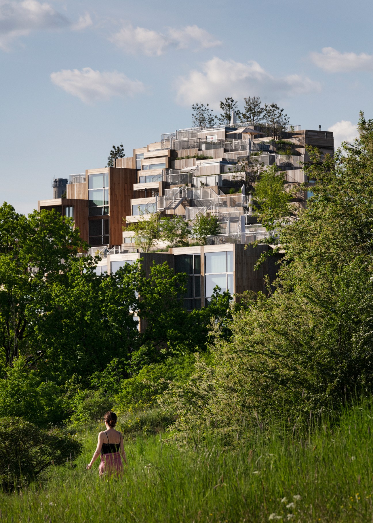 A woman walks through tall grass toward a multi-tiered wooden apartment building with staggered balconies and vegetation.