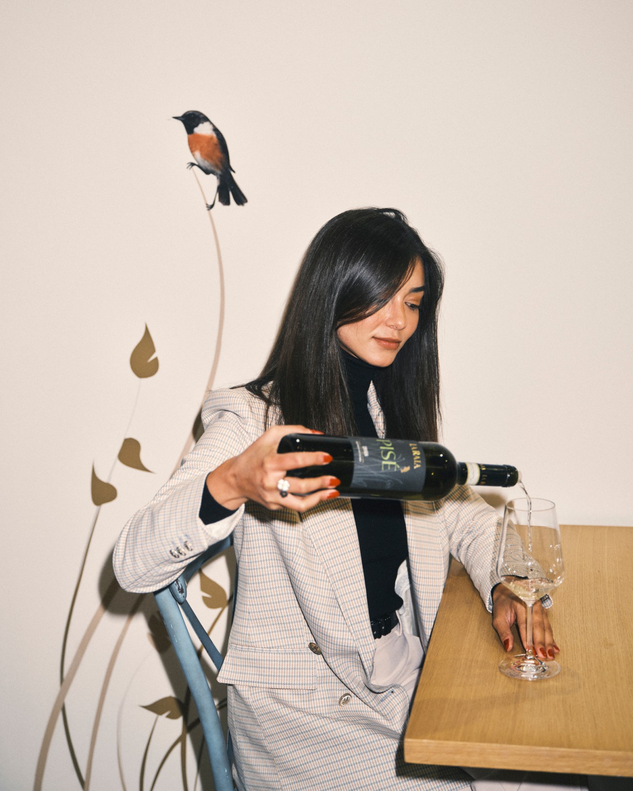 Woman in checkered blazer pouring wine from dark bottle into glass at wooden table