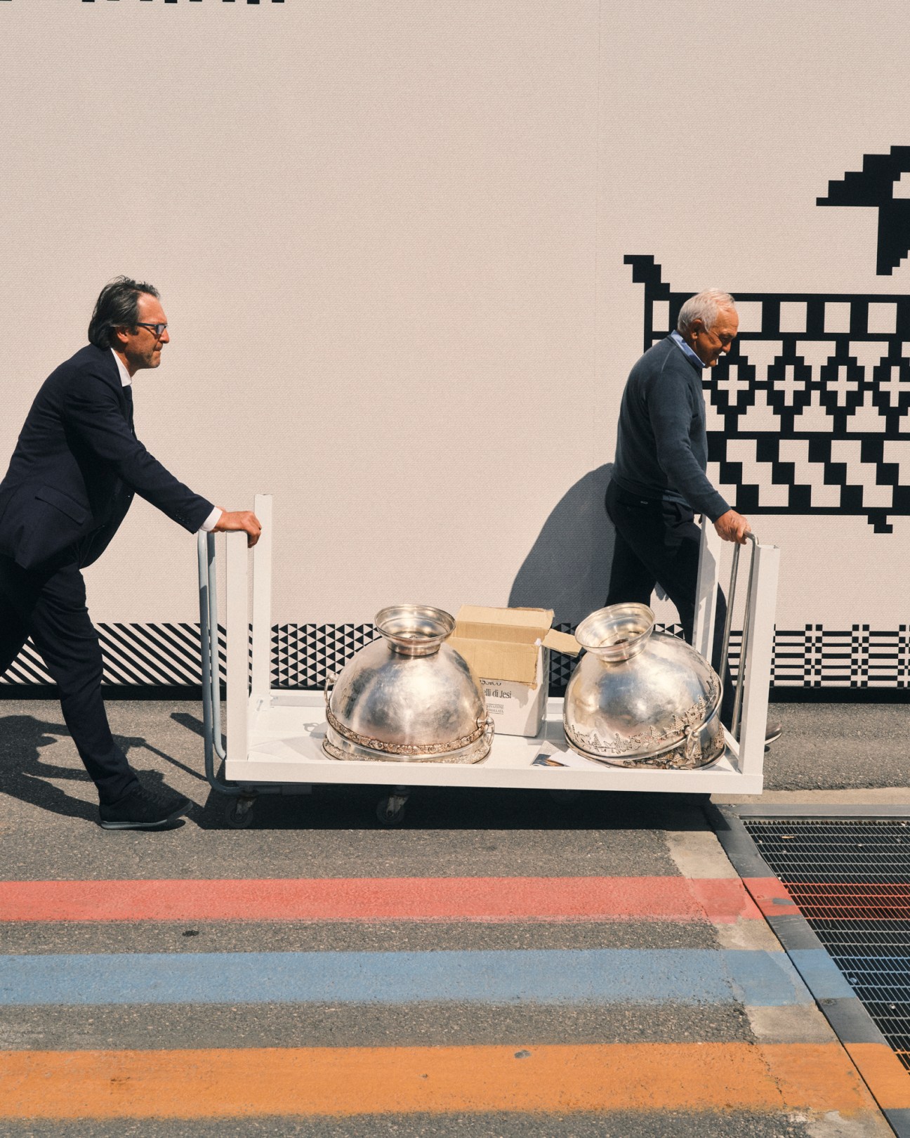 Two men in suits transporting wine equipment on a wheeled cart at Vinitaly trade fair