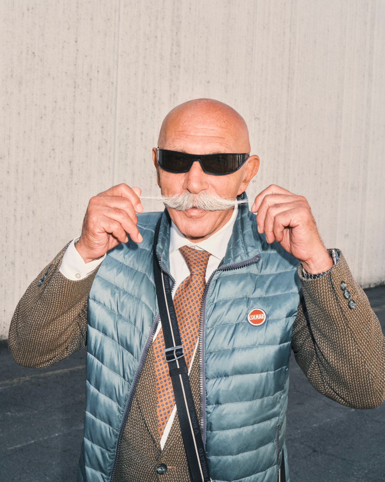 Bald man with impressive white mustache wearing sunglasses and light blue quilted vest at wine fair