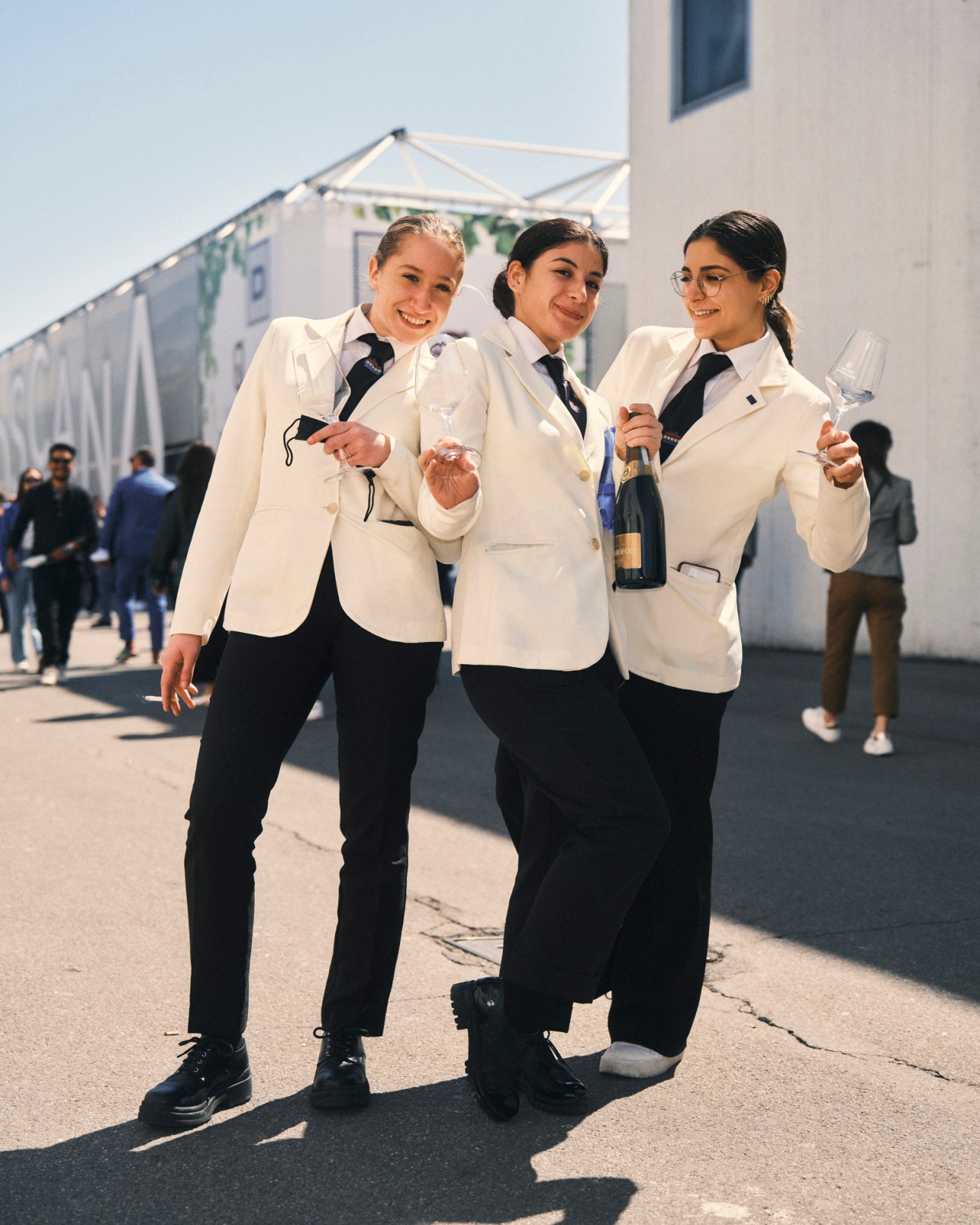 Three young women in matching white blazers and black ties posing together outdoors at Vinitaly wine fair