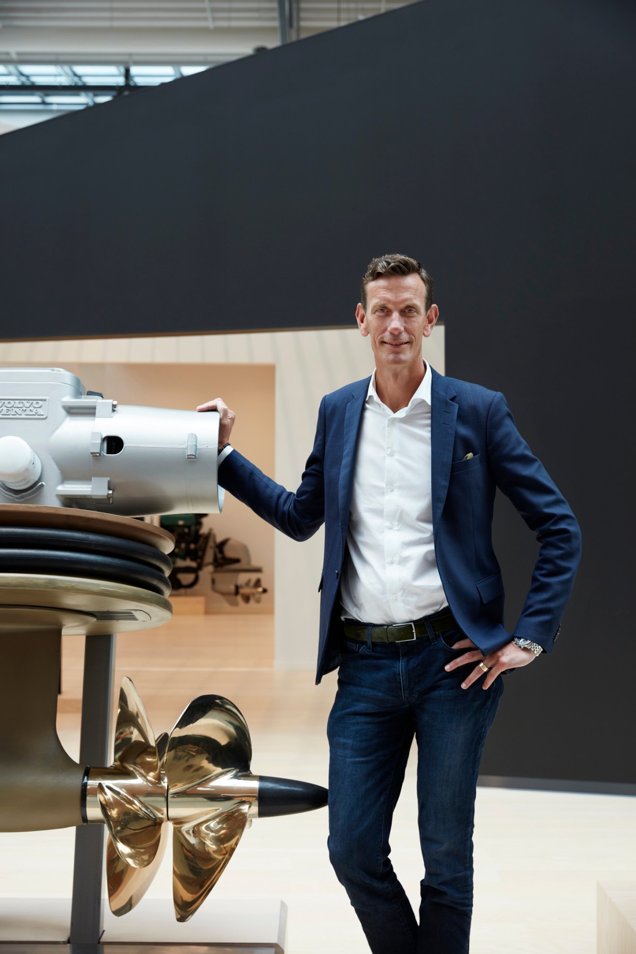 Johan Inden in navy blazer standing next to white marine engine with golden propeller in modern showroom