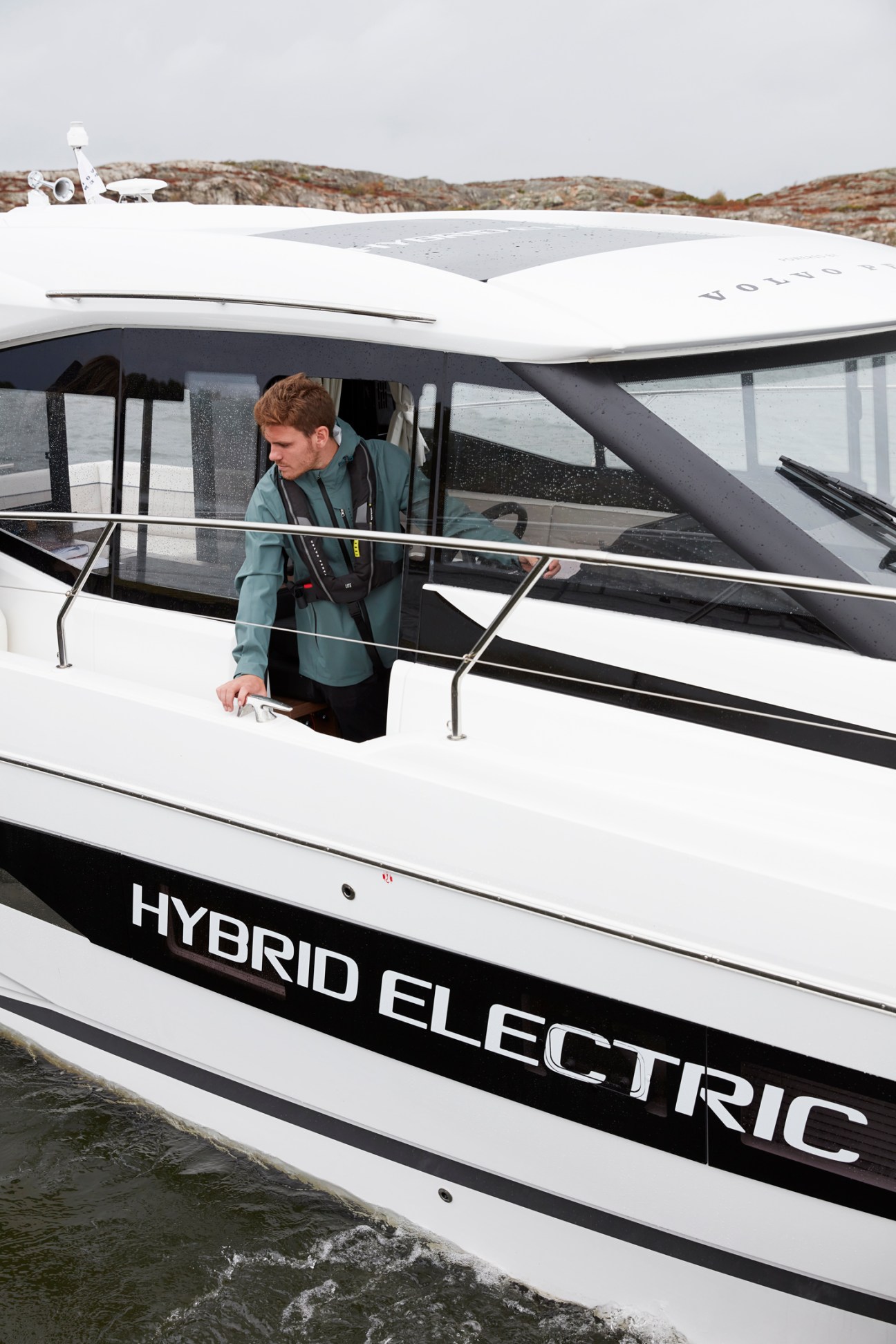 Man in green jacket operating white hybrid electric boat with 'HYBRID ELECTRIC' text on hull at Swedish test facility