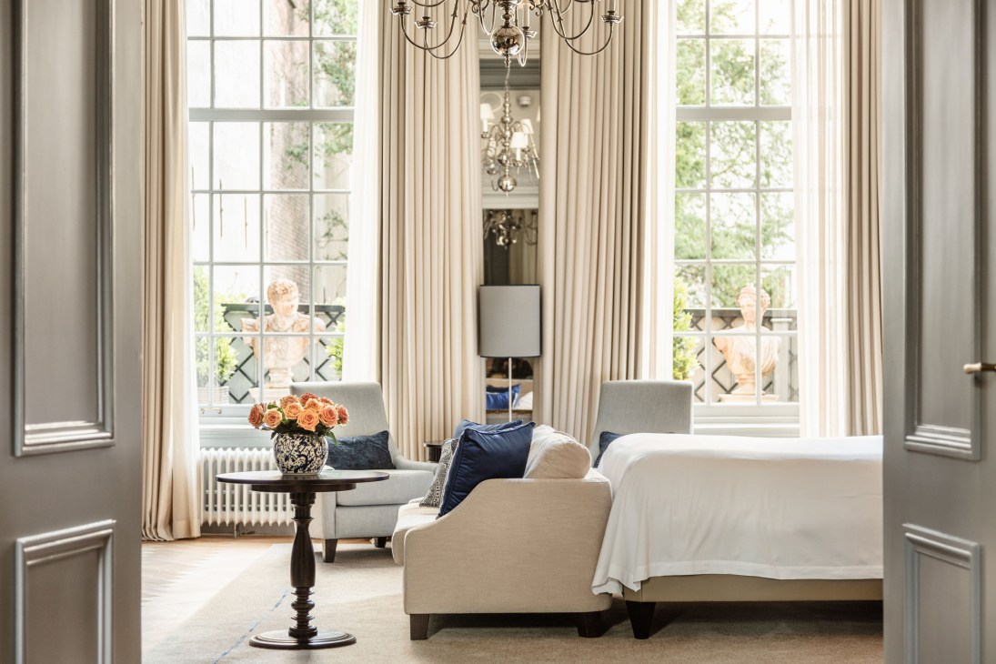 Elegant hotel suite with white sofa, navy pillows, chandelier, tall windows and cream curtains