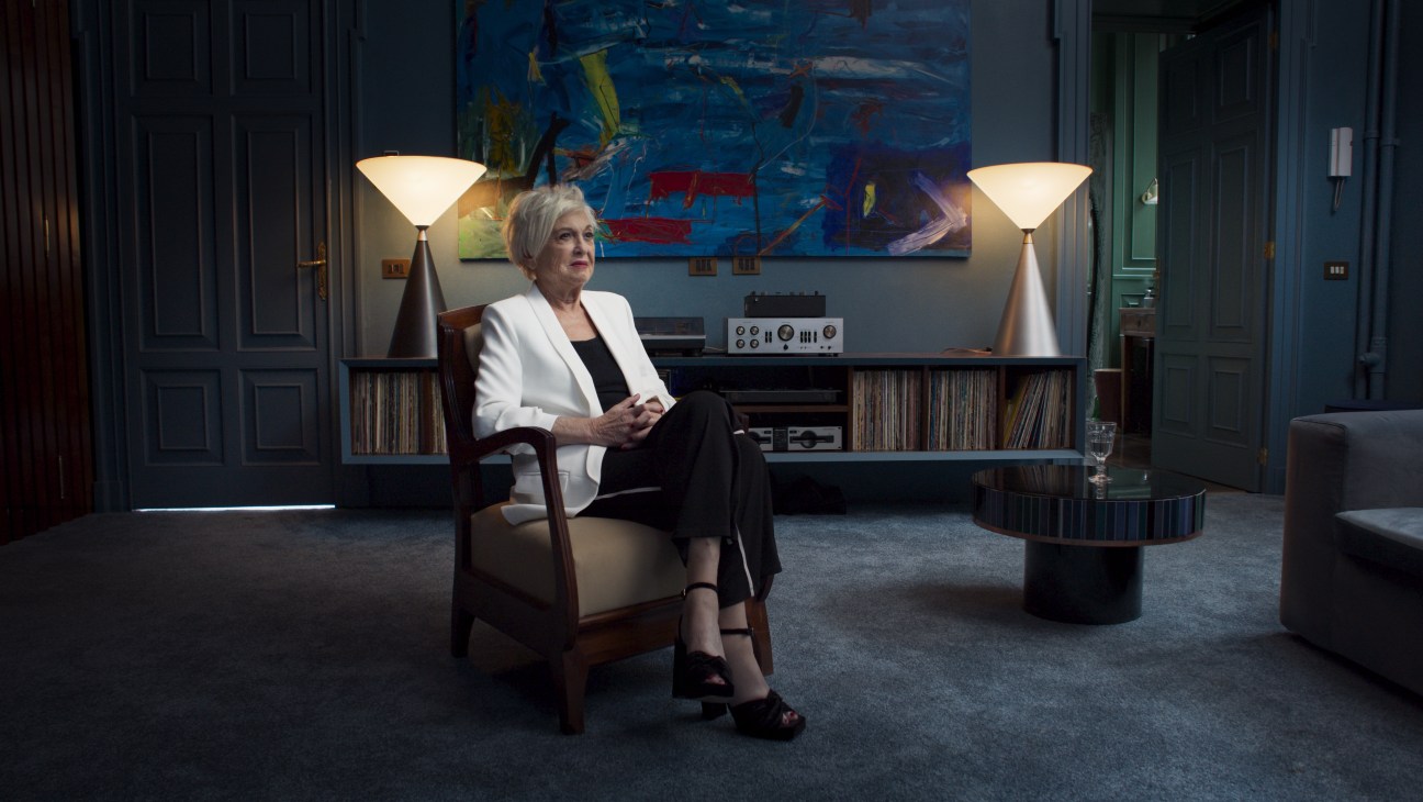 Woman with gray hair in white blazer sitting in elegant room with vinyl records and turntable
