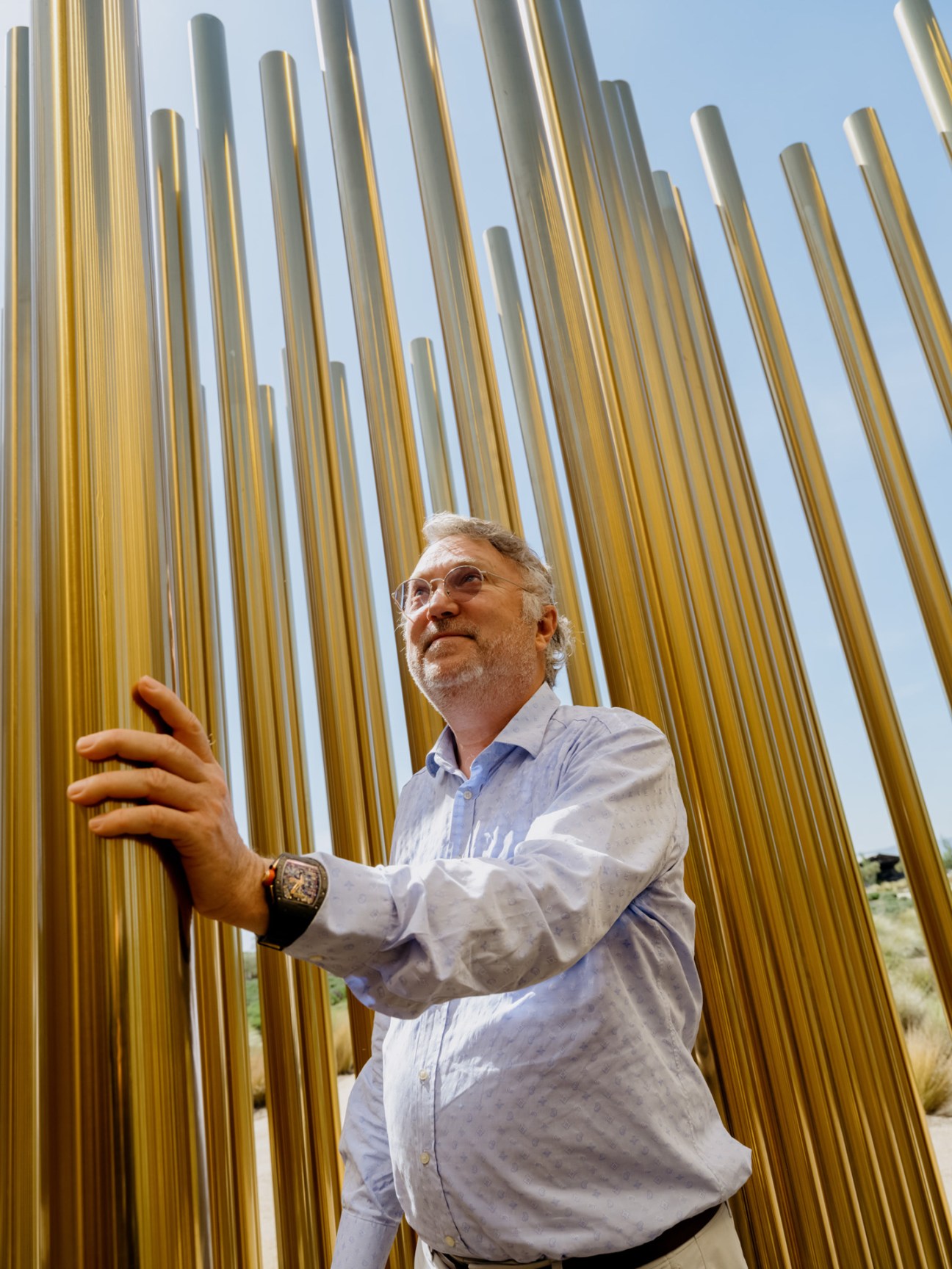 Man in light blue shirt touching golden vertical pipes of large-scale sculpture installation.