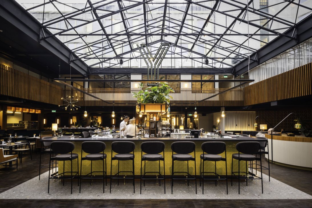 Glass-roofed restaurant at Yalo Urban Boutique Hotel with central bar, black bar stools, wooden paneling, and hanging plants in Ghent