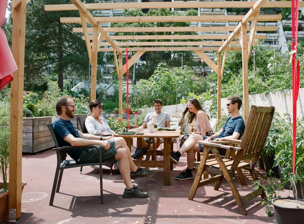 Five people sitting around a wooden table under a pergola in a community garden at Alterlaa housing complex