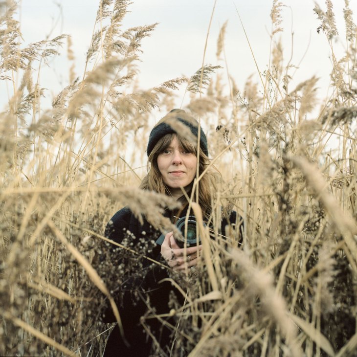 Trisha Ward portrait shot by Zed Nelson outdoors sitting in tall grass wearing dark clothing and beanie
