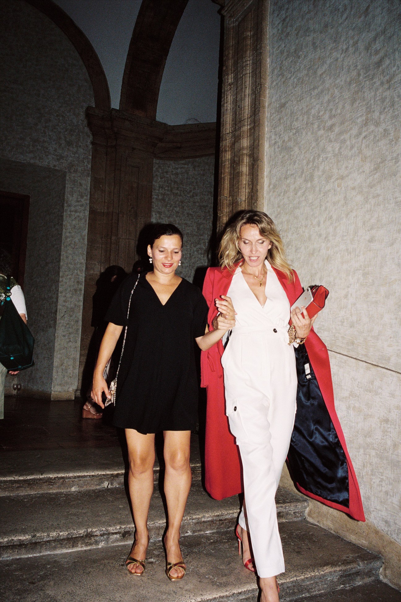 Two elegantly dressed women walking together at a formal embassy reception, one wearing a red coat.