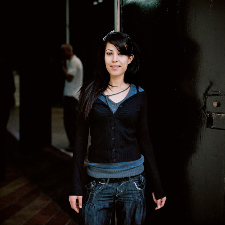 Inès, 22-year-old acrobat at Tunis National School of Circus Arts, standing in a dark corridor wearing a dark cardigan with blue trim.