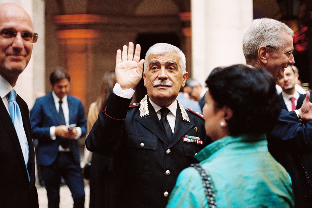 Military official in dress uniform with white hair raising hand in greeting to embassy guests.