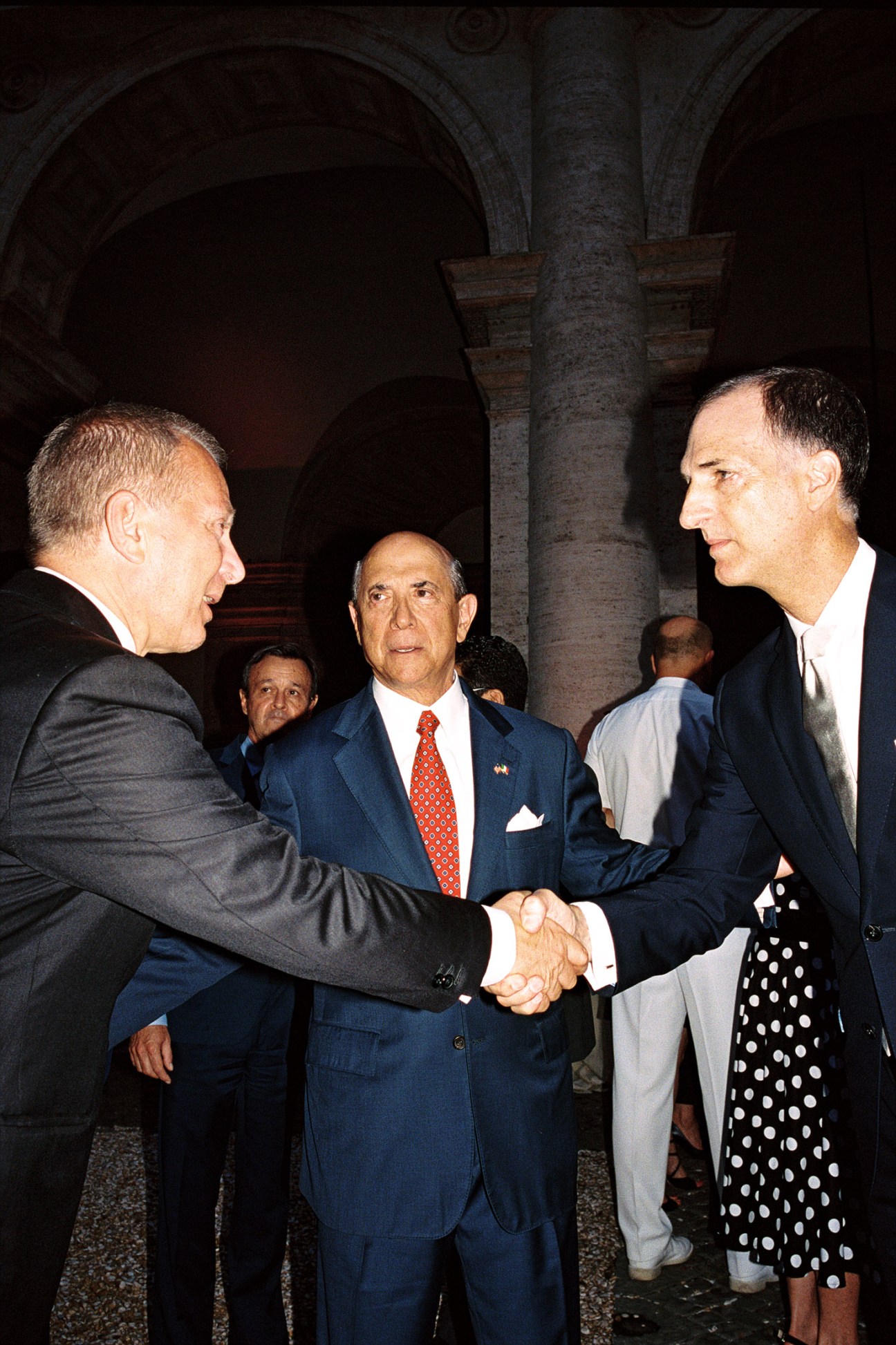 Men in formal suits shaking hands at an embassy reception with Renaissance architecture in background.