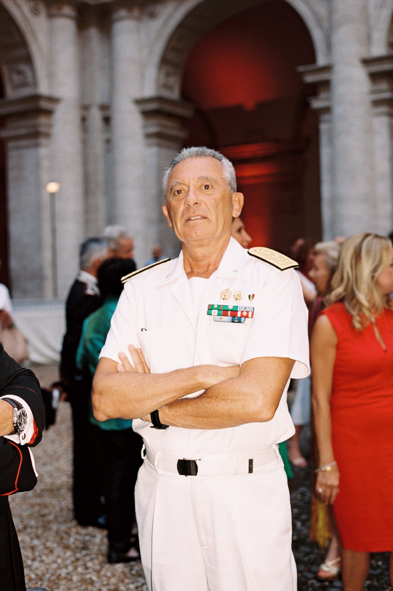 Military officer in white uniform with medals standing in ornate Renaissance corridor at diplomatic reception.