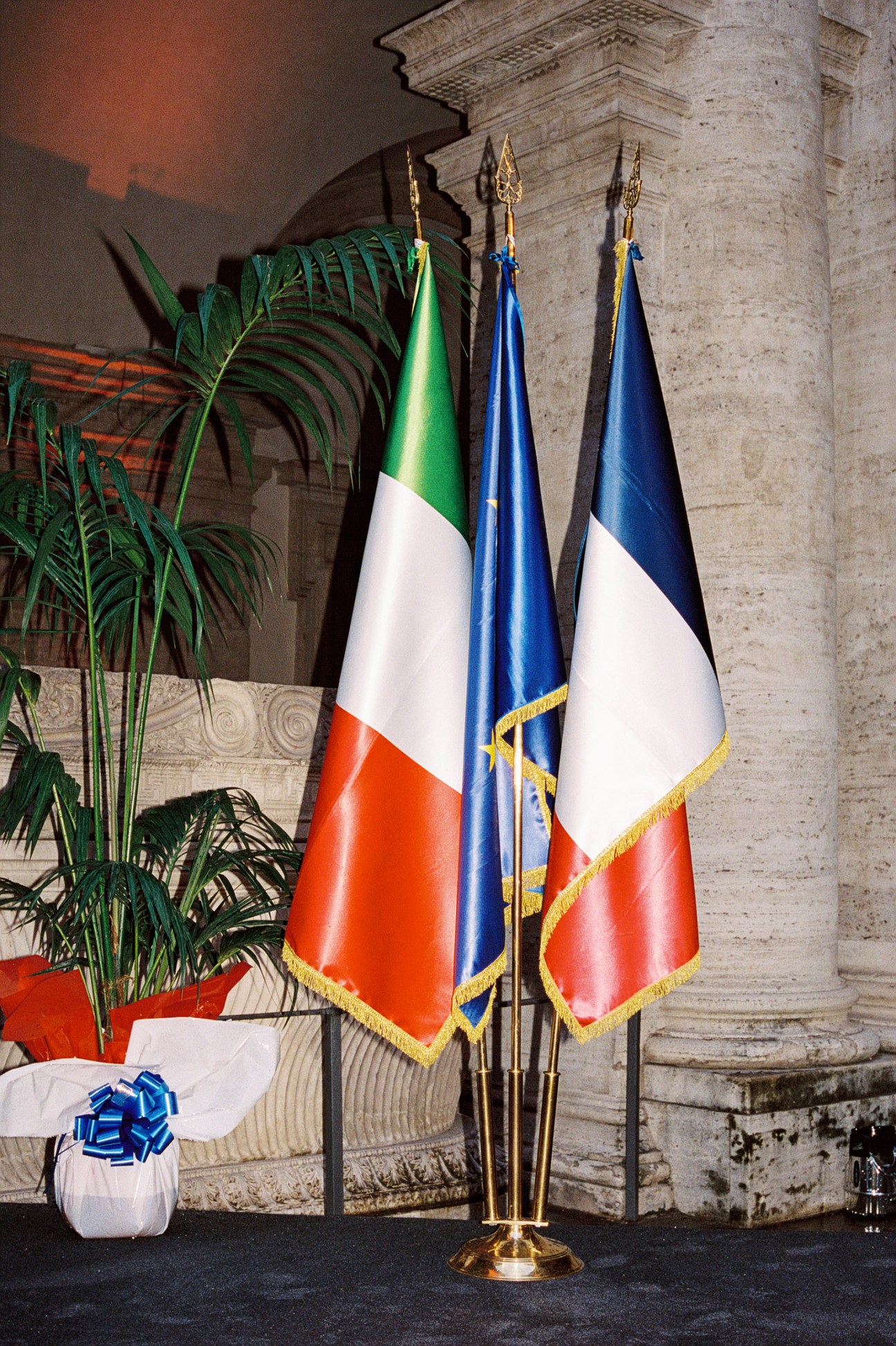 Three flags including the Italian and French flags displayed at a diplomatic event in an ornate stone corridor.