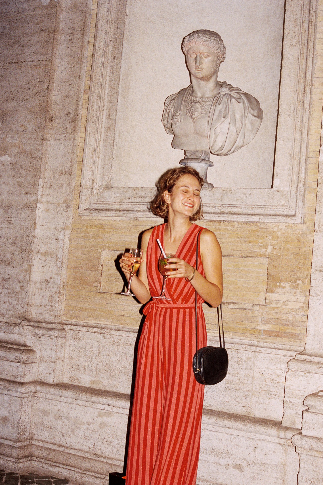 Woman in red striped dress holding cocktail and black handbag in front of classical bust sculpture.