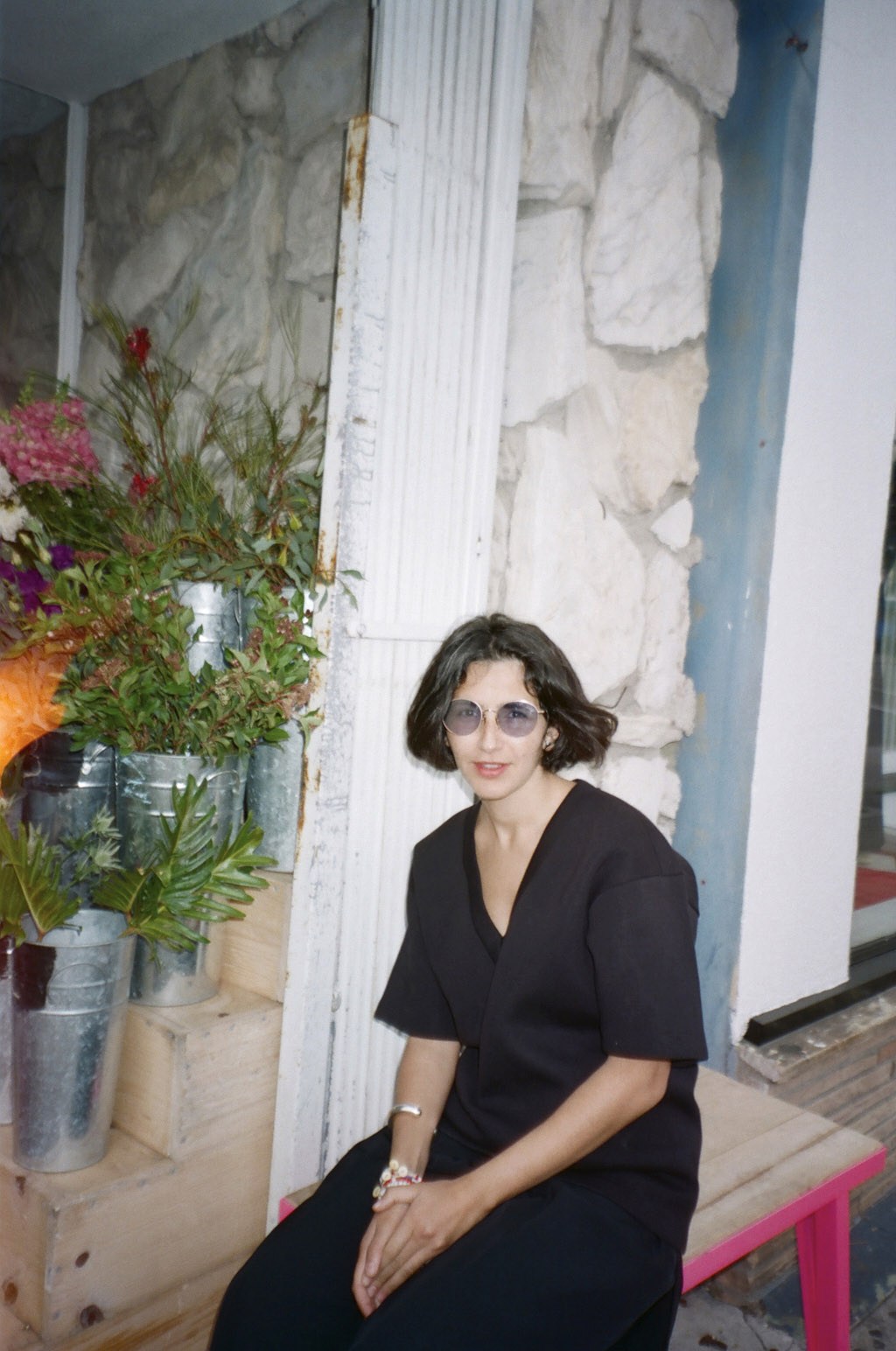 Ahlem Manai-Platt wearing round glasses and black clothing, seated on a pink bench against a white wall with potted flowers.
