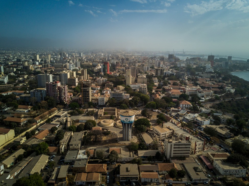 Aerial view of Dakar showing urban sprawl with mixed-height buildings, a water tower, and the Atlantic Ocean coastline