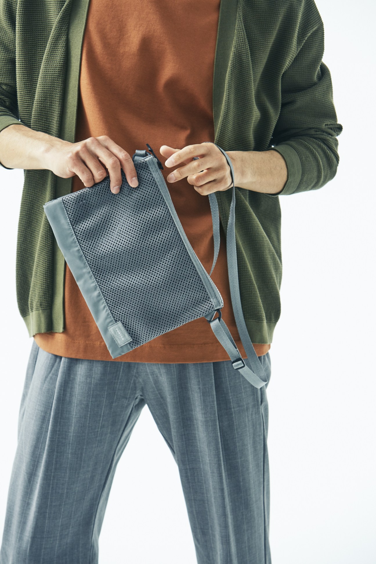 Man holding blue Porter bag wearing green cardigan, brown t-shirt and grey trousers