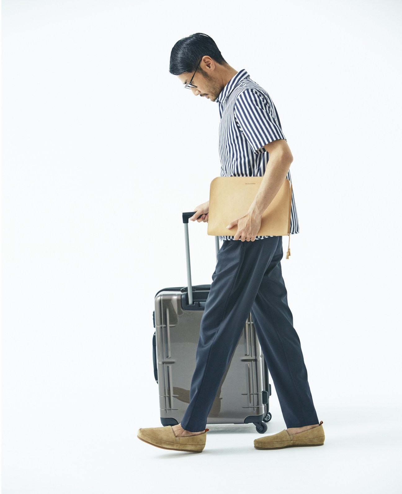 Man in striped shirt and trousers holding tan portfolio case while pulling a grey suitcase.