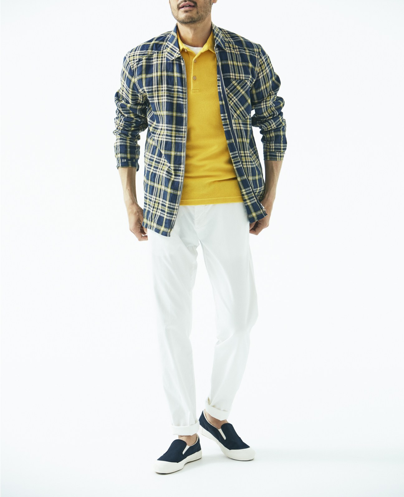 Man wearing plaid jacket, yellow polo shirt, white trousers and navy slip-on trainers against white background.