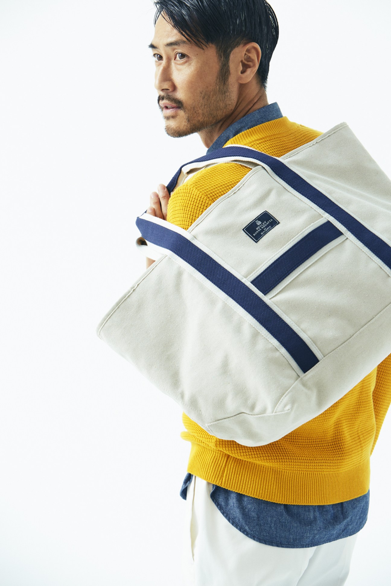 Man wearing yellow jumper and denim shirt carrying cream canvas backpack with navy straps