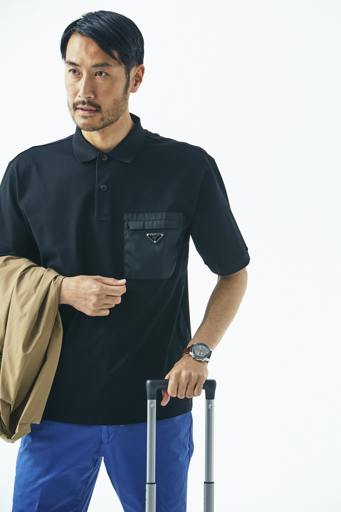 Man wearing black Prada shirt, Descente Pause jacket, blue Tomorrowland shorts, and Omega watch holding Victorinox suitcase