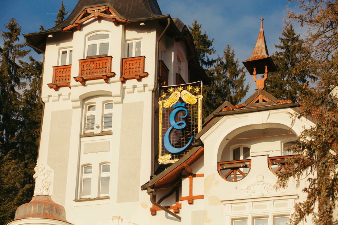 A white villa with ornate balconies and a church spire in the background among trees
