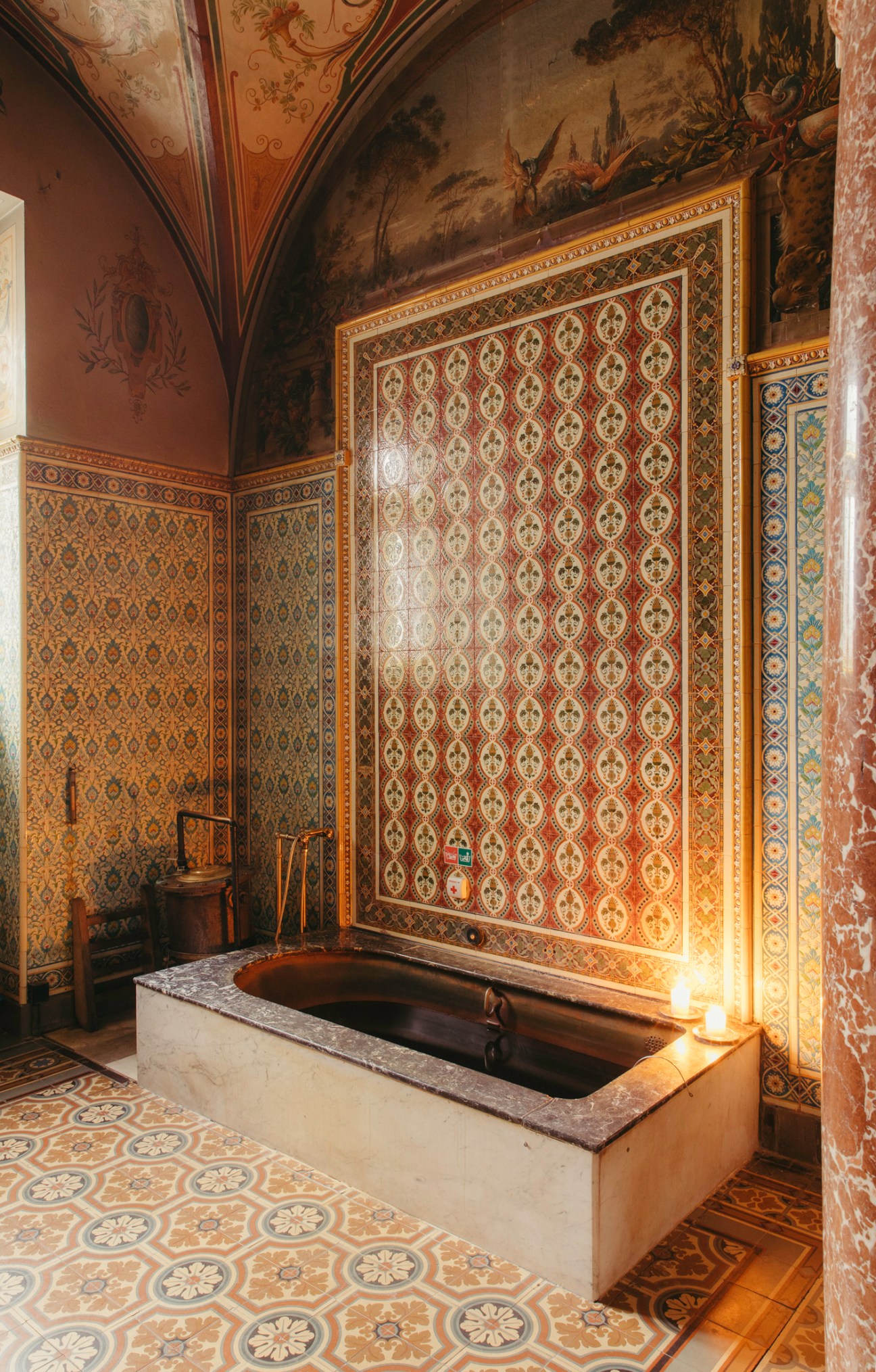 Ornate bathroom with decorative tiled walls, vaulted ceiling, and sunken mineral bath in historic spa treatment room