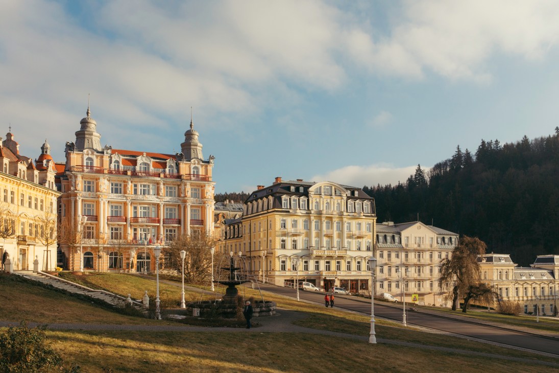 Historic spa buildings with ornate facades and towers line a tree-lined street in Marienbad