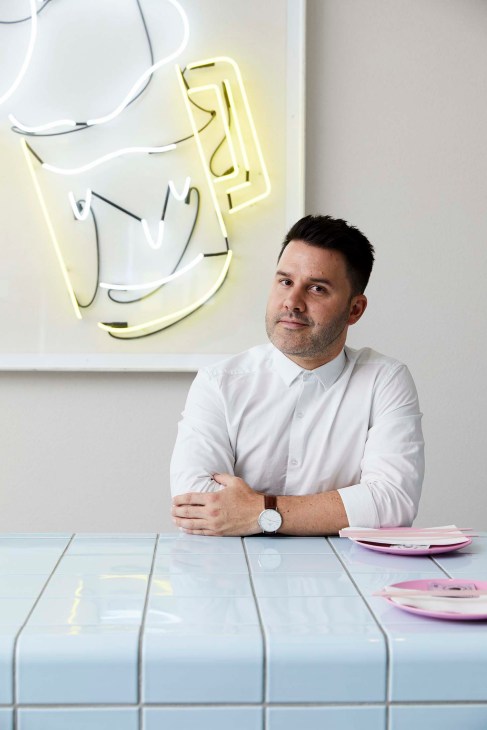 Interior designer Ryan Genesin sitting at a table with neon artwork on the wall behind him.